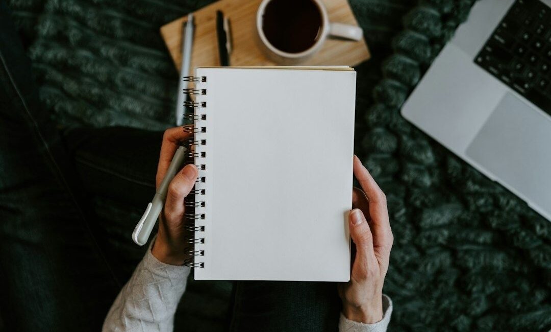 Hænder holder en blank notesbog med pen, kaffe og bærbar computer